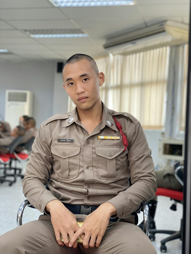 A Thai police officer sits indoors wearing uniform and looking serious.